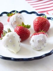 A plate of fresh strawberries, some of which are coated in white chocolate and adorned with small star decorations. The plate has a scalloped edge and there is a red, white, and blue striped cloth in the background, suggesting a patriotic theme.