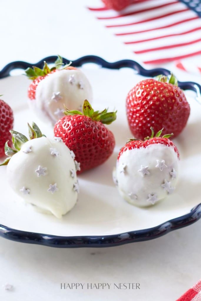 A plate of fresh strawberries, some of which are coated in white chocolate and adorned with small star decorations. The plate has a scalloped edge and there is a red, white, and blue striped cloth in the background, suggesting a patriotic theme.