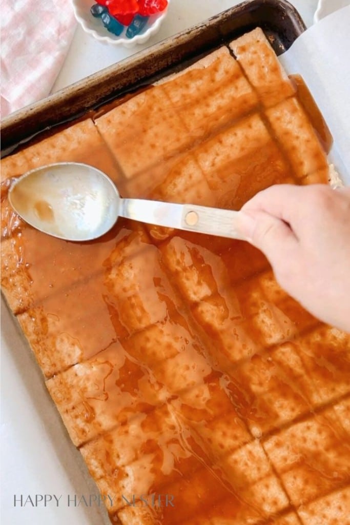 A hand uses a spoon to drizzle syrup over a large sheet of matzo placed on a baking tray. The matzo is scored into a grid pattern. In the background, there is a small bowl containing colorful gummy candies.
