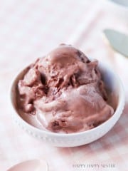 A close-up of a bowl filled with creamy chocolate ice cream, placed on a soft, light pink checkered cloth. There's a blurred spoon and knife in the background. The texture of the ice cream appears smooth and slightly melty.