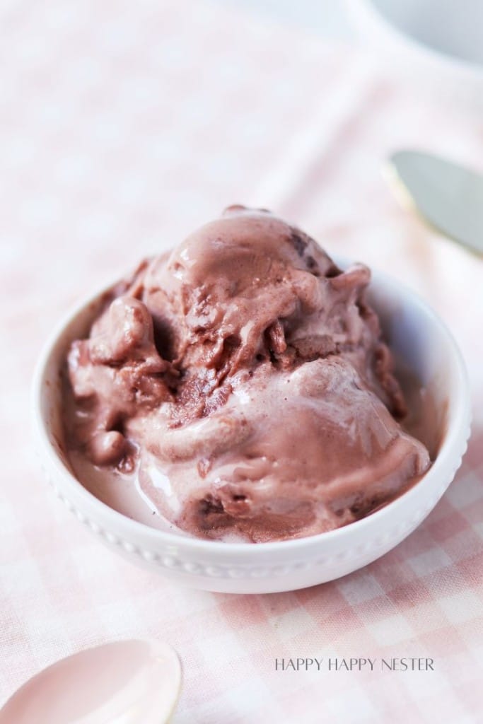 A close-up of a bowl filled with creamy chocolate ice cream, placed on a soft, light pink checkered cloth. There's a blurred spoon and knife in the background. The texture of the ice cream appears smooth and slightly melty.