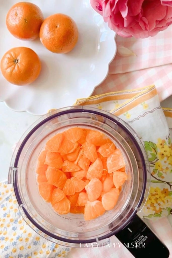 Top-down view of three whole oranges on a white plate, a bowl filled with orange segments, and floral-patterned cloths beside them. There are also some pink flowers visible in the top right corner.