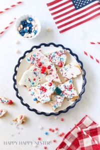 A plate of festive patriotic bark on a decorative plate, topped with red, white, and blue sprinkles and candy. Surrounding the plate are scattered candies, a small bowl of colored candies, red striped straws, an American flag, and a red gingham napkin.
