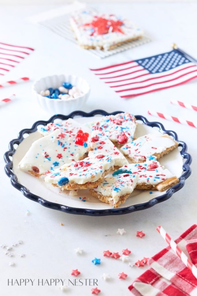 A plate filled with star-shaped cookies covered in white frosting and red, white, and blue sprinkles. Surrounding the plate are small American flags, a bowl of M&Ms, red and white paper straws, and a red checkered napkin, evoking a festive, patriotic theme.