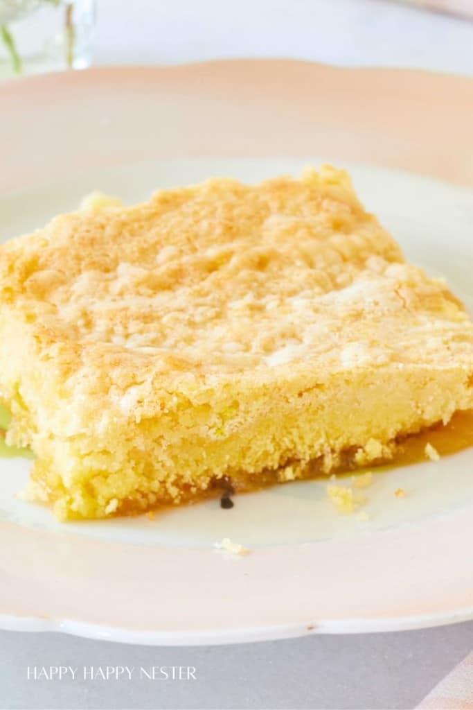 A slice of yellow cake with a crumbly, golden-brown top sits on a white plate with a scalloped edge. The background is blurred, highlighting the cake piece prominently. The text "Happy Happy Nester" is visible at the bottom of the image.