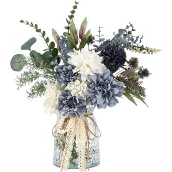 A bouquet of artificial flowers in shades of blue, white, and green is arranged in a glass jar. The jar is decorated with a beige lace ribbon tied in a bow. The arrangement includes flowers like dahlias and greenery such as eucalyptus.