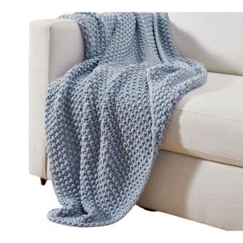 A chunky knit gray throw blanket is draped over the arm of a light-colored sofa. The blanket has a textured, thick weave, contributing to a cozy and inviting atmosphere in the room.