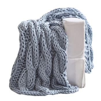 A chunky knit blanket in light grey-blue color is draped over a white upholstered chair. The thick, braided pattern of the blanket creates a cozy and textured appearance, perfect for adding warmth and style to a living space.