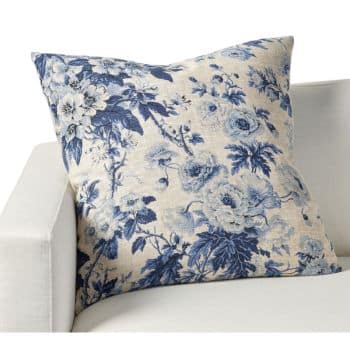 A square throw pillow with a blue floral pattern on a beige background is placed on a white upholstered chair. The design features various blooming flowers and leaves in a vintage style. The pillow is plump and adds a touch of elegance to the chair.