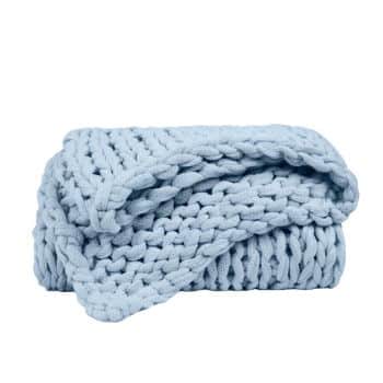 A neatly folded, chunky knit blanket in a soft pastel blue color. The blanket features thick, large stitches that give it a cozy and plush appearance. It is positioned against a plain white background.