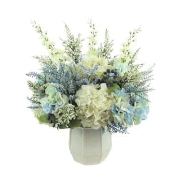 A white vase holds a bouquet of artificial flowers, featuring light blue, white, and green hydrangeas, interspersed with delicate, feathery greenery and small blue accents. The arrangement has a fresh and elegant appearance.