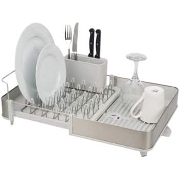 A dish rack holds plates, a wine glass, utensils, and a mug. The rack features vertical prongs for the plates and a slotted tray for utensils. It also has a drainage board underneath for excessive water to be collected. The dish rack is minimalist and modern in design.