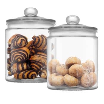 Two glass jars with glass lids sit side by side. The jar on the left contains striped, crescent-shaped pastries, while the jar on the right holds round, powdered sugar-coated pastries. Both jars are transparent, clearly displaying the treats inside.