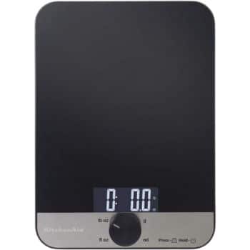 A digital kitchen scale with a black surface and a circular control knob at the bottom center. The display screen shows "0.0" and has buttons for different units of measurement. The brand name "KitchenAid" is visible on the lower left part of the scale.