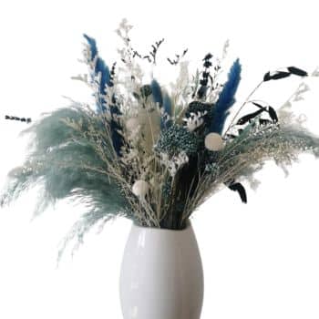 A decorative arrangement of dried flowers and foliage in a white vase. The bouquet features a mix of blue, green, and white plants, including pampas grass and delicate stems, creating a textured and colorful display against a plain background.