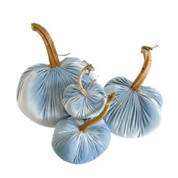 Four light blue velvet pumpkins with long, curved stems arranged together. The fabric texture and soft color give a plush and elegant appearance to the decorative items. The sizes vary, creating a visually appealing contrast.