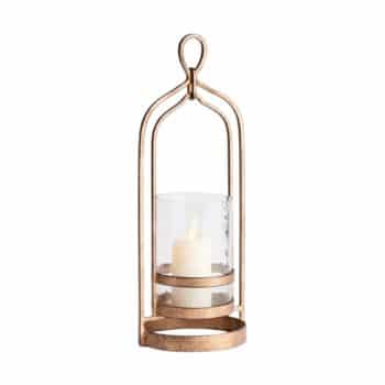 A metallic lantern with a gold finish holding a lit white candle inside a clear cylindrical glass holder. The lantern has an arched frame with a loop at the top for hanging or carrying. The design is elegant and vintage-inspired.