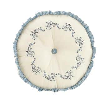 A round, white cushion adorned with intricate blue floral embroidery in a circular pattern. The edges are trimmed with blue ruffled fabric, and there is a central blue button tuft.