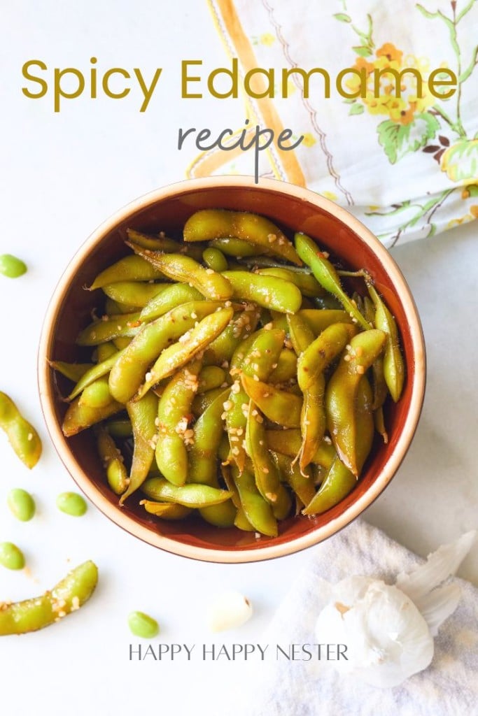 A bowl of spicy edamame garnished with sesame seeds is placed on a white surface. Nearby, edamame pods, a garlic bulb, and a decorative cloth featuring yellow flowers are visible. The text "Spicy Edamame recipe" is at the top, with "HAPPY HAPPY NESTER" at the bottom.