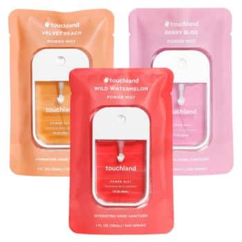 Three Touchland Power Mist hand sanitizers are displayed in their packaging. The scents are Velvet Peach (orange packaging), Wild Watermelon (red packaging), and Berry Bliss (pink packaging). Each package contains a compact, white dispenser with colored mist inside.