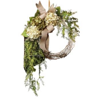 A partially decorated wreath featuring light-colored hydrangea flowers, a large beige burlap bow, and green leafy vines that drape down from one side. The wreath has a natural, rustic appearance with a mix of textures.