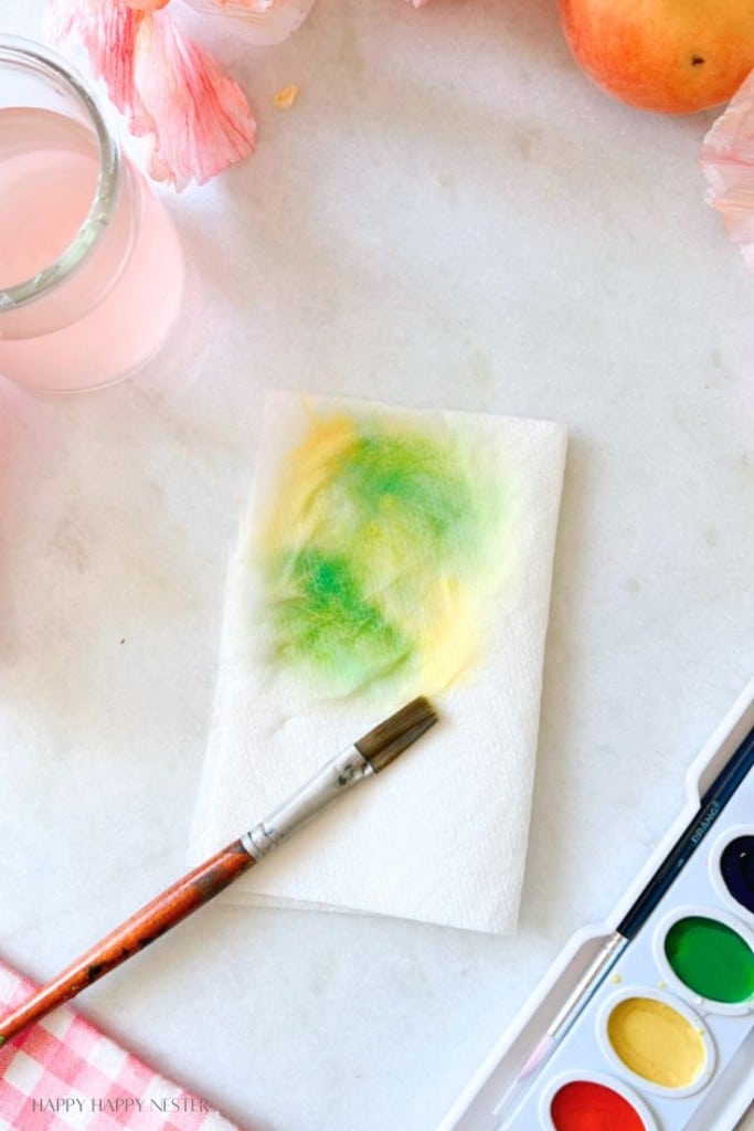 A paintbrush rests on a napkin with green and yellow watercolor stains beside a watercolor paint set, a glass of pink-tinted water, and a peach on a white surface with pink flowers in the background.