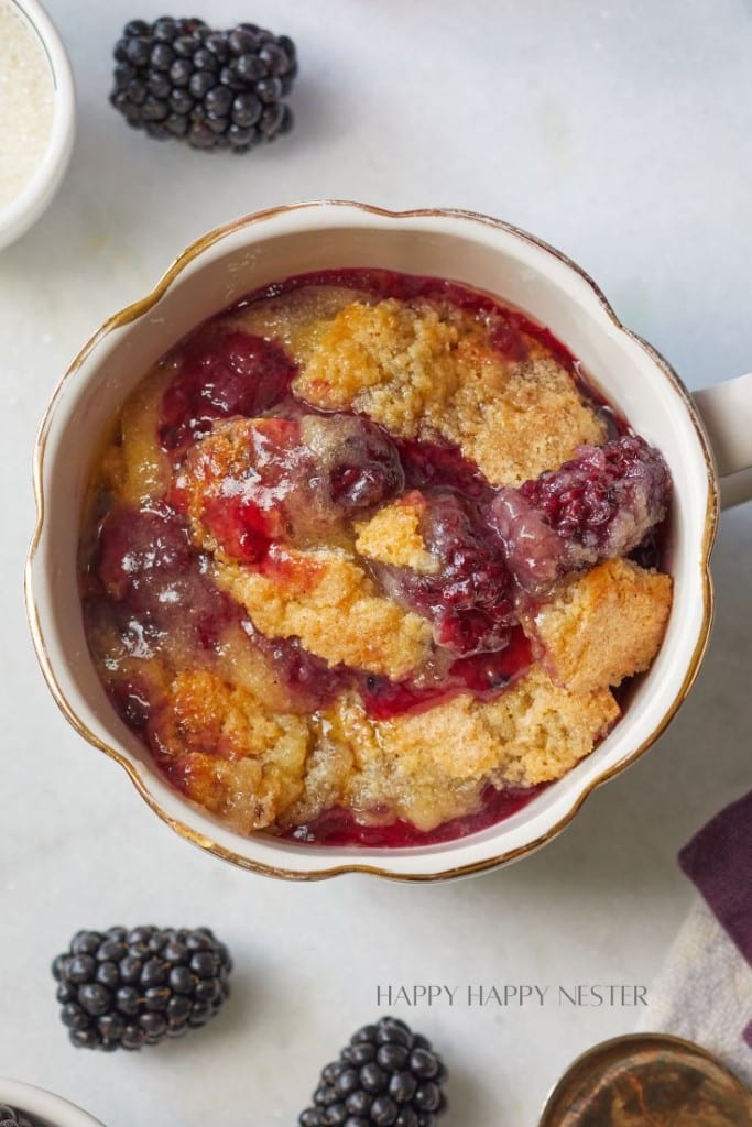 A close-up of a ceramic dish filled with blackberry cobbler. The cobbler features a golden, crumbly topping with vibrant purple-black blackberry filling peeking through. Fresh blackberries are scattered around the dish on a marble surface.