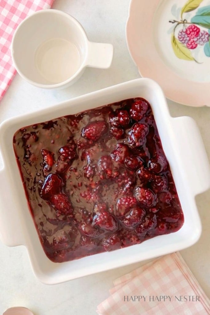 A white ceramic baking dish filled with dark red berry compote placed on a marble surface. Next to it are a white mug, a decorative plate with a fruit design, and a pink and white gingham cloth. The image has the text "HAPPY HAPPY NESTER" at the bottom right.