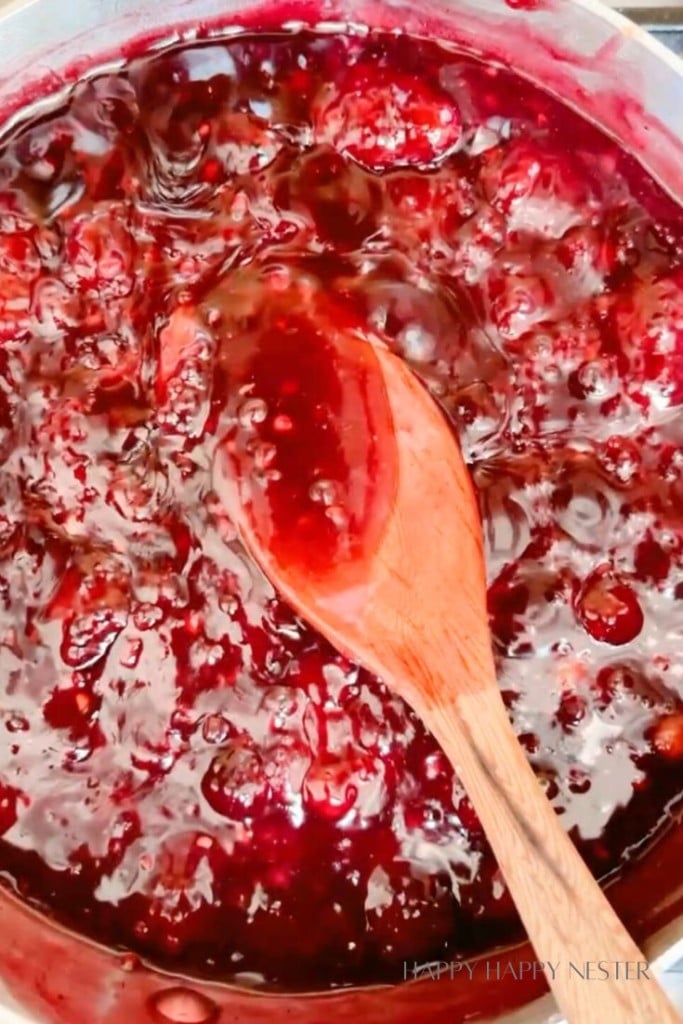 A wooden spoon stirring a pot full of rich, bubbling, dark red cranberry sauce with visible cranberry chunks. The glossy texture and deep color indicate the sauce is thickening as it cooks.