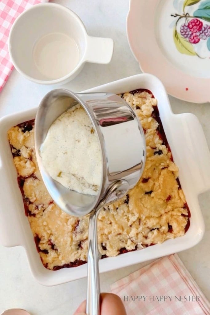 A hand holds a saucepan, pouring a creamy mixture over a baked dessert in a white square dish. Nearby are an empty ceramic bowl and a pink and white checked cloth. A floral-patterned plate is partly visible in the background.