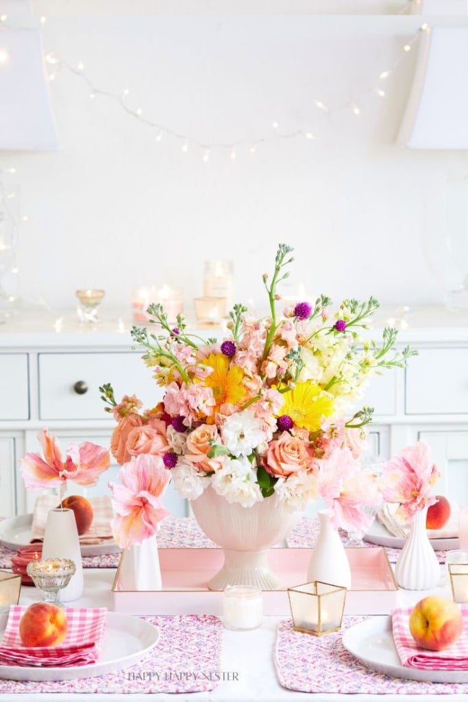 A beautifully set dining table with a centerpiece of vibrant flowers in shades of pink, yellow, white, and green. The table features a pink and white color scheme with patterned placemats, candles, and decorative plates with peaches. Fairy lights hang in the background.