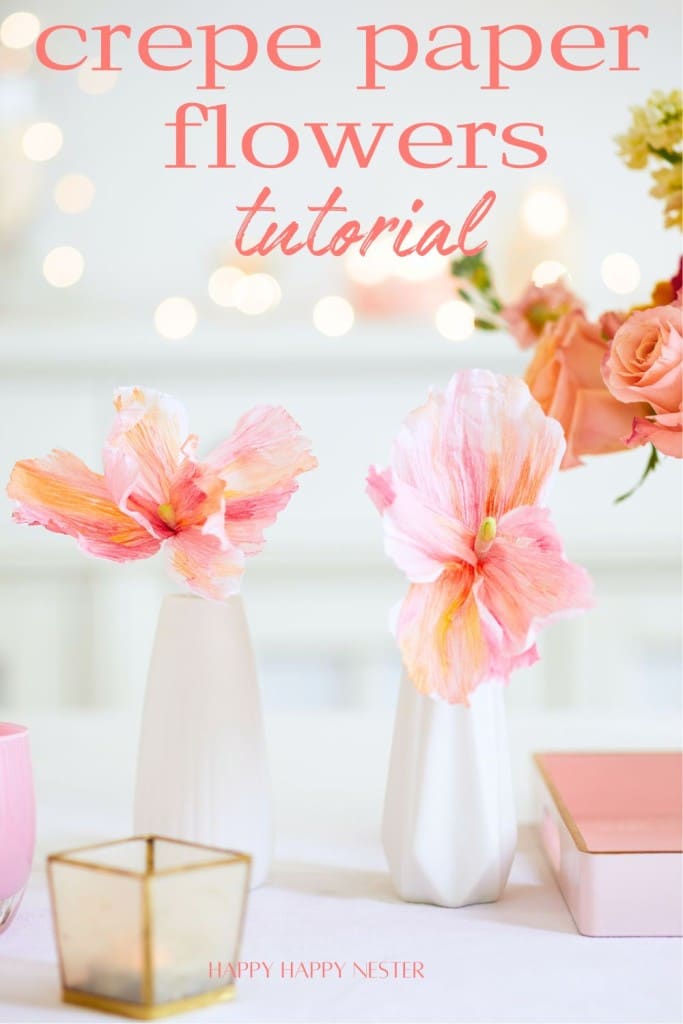 A delicate arrangement of crepe paper flowers in pastel pinks and oranges is displayed in white vases against a soft, blurred background. Text at the top says "crepe paper flowers tutorial." A small golden candle holder and a light pink notebook are also visible.