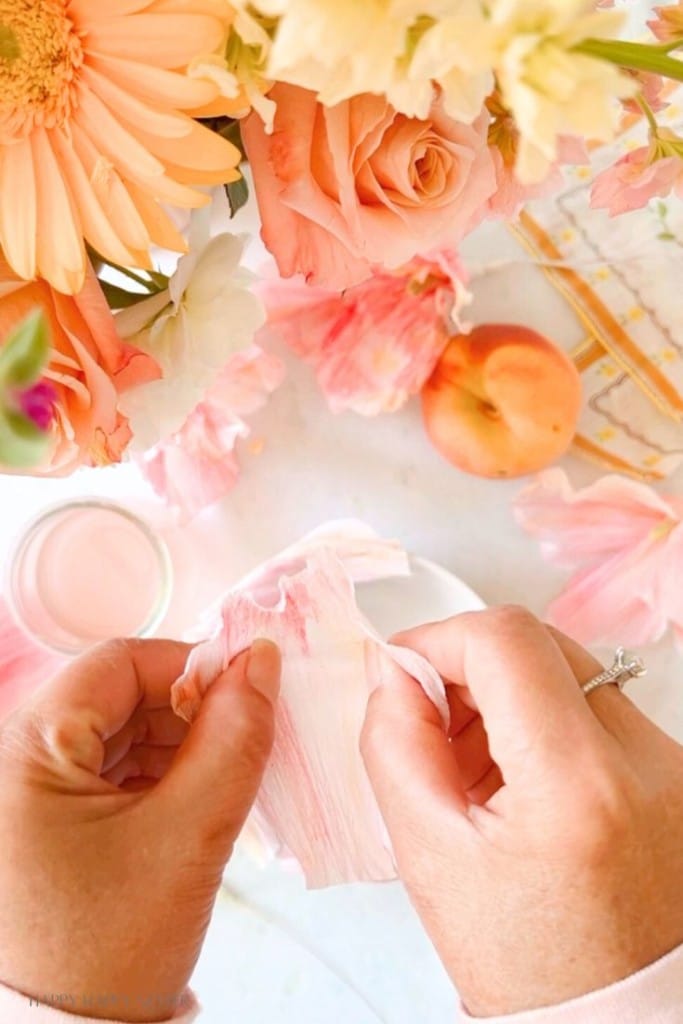 A person with a ring on their left hand is seen peeling a pink and white petal. The scene is surrounded by various colorful flowers, a peach, and a glass of a light pink drink on a white surface. The setting appears bright and vibrant.