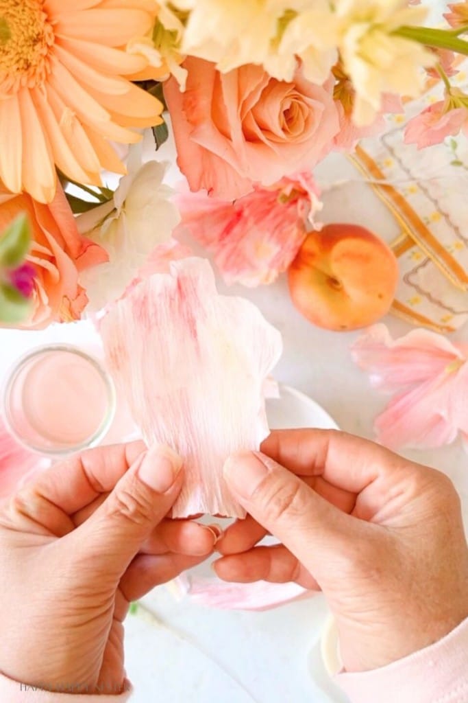 A pair of hands holding a light pink flower petal over a table adorned with various flowers, including roses and daisies, as well as a peach. A small glass with a pink liquid is also visible on the table.