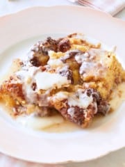 A close-up of a plate with a serving of bread pudding topped with a generous drizzle of white icing. The bread pudding has a golden-brown crust with visible chunks of bread and a creamy interior. The plate rests on a pink and white striped cloth.