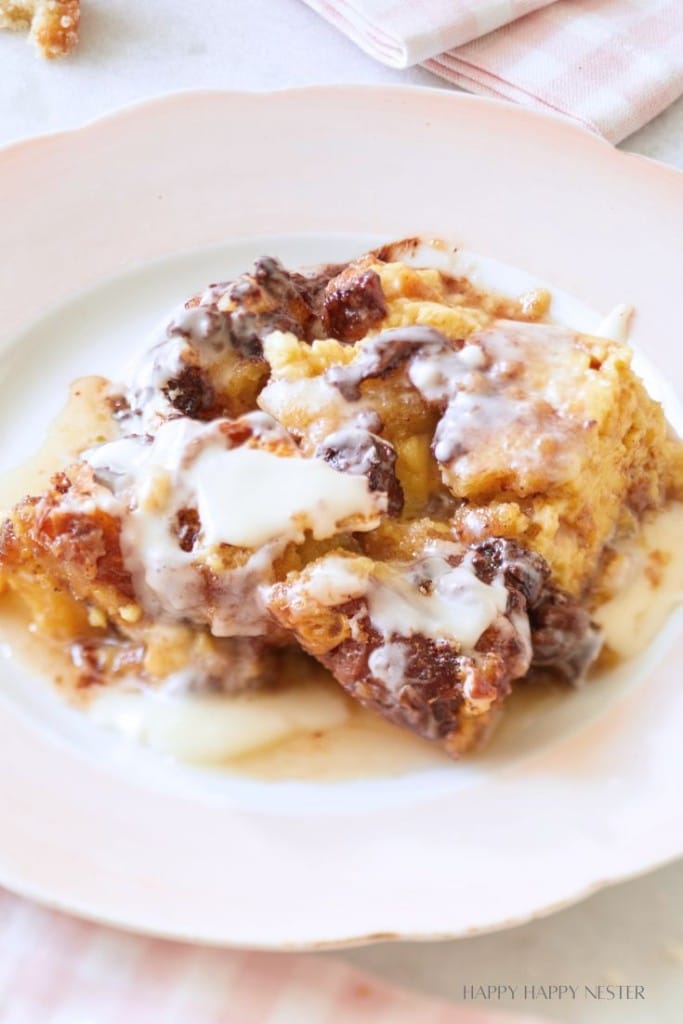 A close-up of a plate with a serving of bread pudding topped with a generous drizzle of white icing. The bread pudding has a golden-brown crust with visible chunks of bread and a creamy interior. The plate rests on a pink and white striped cloth.