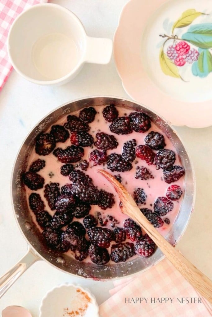 A saucepan filled with blackberries mixed in a creamy, pink liquid, with a wooden spoon stirring the contents. Nearby are a white cup, a decorative plate with a floral design, a small dish of cinnamon, and a pink checkered cloth. The text "Happy Happy Nester" is in the corner.