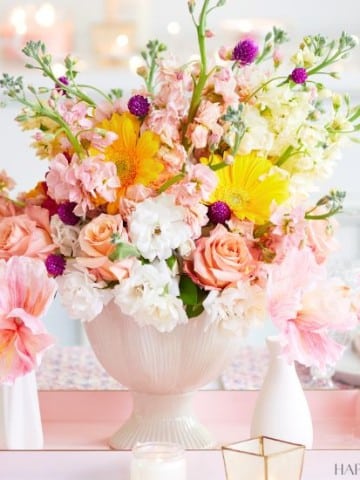 A vibrant floral arrangement in a white vase sits on a table, surrounded by pink and white candles. The bouquet features a mix of pink roses, yellow gerbera daisies, white stock flowers, and small purple blooms. The background is softly blurred, with a hint of a cozy setting.