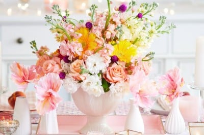 A vibrant floral arrangement in a white vase sits on a table, surrounded by pink and white candles. The bouquet features a mix of pink roses, yellow gerbera daisies, white stock flowers, and small purple blooms. The background is softly blurred, with a hint of a cozy setting.