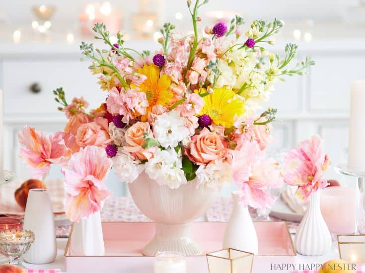 A vibrant floral arrangement in a white vase sits on a table, surrounded by pink and white candles. The bouquet features a mix of pink roses, yellow gerbera daisies, white stock flowers, and small purple blooms. The background is softly blurred, with a hint of a cozy setting.