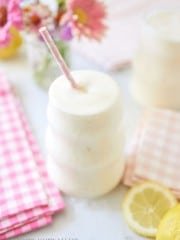 A creamy, frothy drink in a uniquely-shaped glass with a pink straw. The drink is surrounded by pink gingham napkins, fresh lemons, and a vase of colorful flowers. The background is softly blurred, creating a light and airy ambiance.