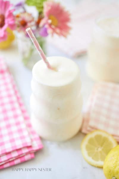 A creamy, frothy drink in a uniquely-shaped glass with a pink straw. The drink is surrounded by pink gingham napkins, fresh lemons, and a vase of colorful flowers. The background is softly blurred, creating a light and airy ambiance.