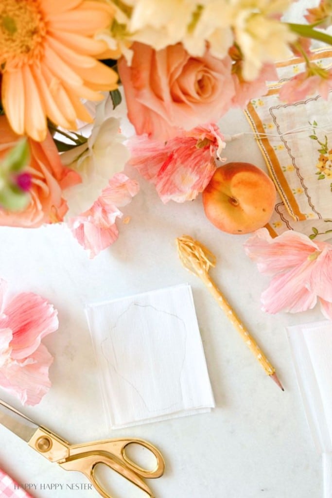 A workspace with peach-colored roses, a daisy, and other flowers. A peach, a gold pen, a paper with a leaf sketch, and scissors are arranged on a white surface. Pink flower petals are scattered around, adding to the colorful and creative atmosphere.