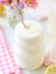 A creamy, frothy drink served in a unique, wavy glass with a pink and white polka-dot straw. The background features a soft-focus arrangement of colorful flowers, a pink checkered napkin, and a sliced lemon.