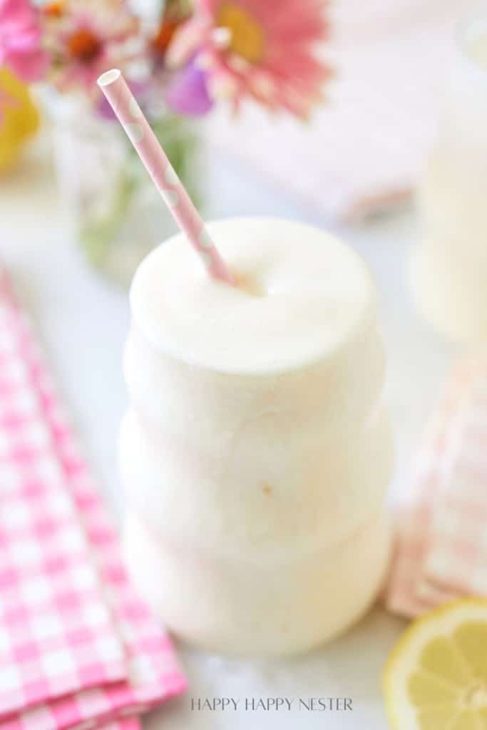 A creamy, frothy drink served in a unique, wavy glass with a pink and white polka-dot straw. The background features a soft-focus arrangement of colorful flowers, a pink checkered napkin, and a sliced lemon.