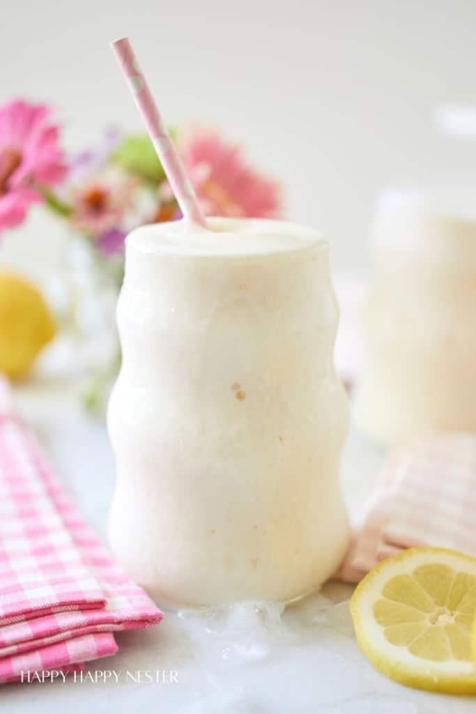 A frothy, creamy drink in a wavy glass with a pink and white polka-dotted straw. The drink is set on a white surface with a pink checkered napkin and a lemon slice nearby. There are blurred pink and white flowers in the background.