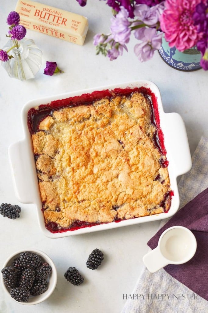 A square white baking dish contains a freshly-baked blackberry cobbler, with a golden, crumbly topping. Surrounding the dish are fresh blackberries, a small pitcher of cream, a butter pack, and a vase with purple flowers, set atop a light-colored surface.