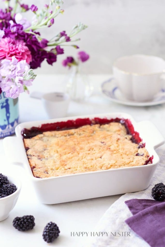 A freshly baked blackberry cobbler in a white square baking dish is placed on a table. There are fresh blackberries scattered around. The table is adorned with a bouquet of purple and pink flowers in the background and a white teacup and saucer beside the dish.