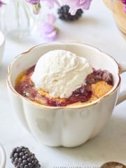 A cup of warm berry cobbler topped with a scoop of vanilla ice cream sits on a white surface. Fresh blackberries and a spoon are nearby, and purple flowers in the background add a decorative touch.