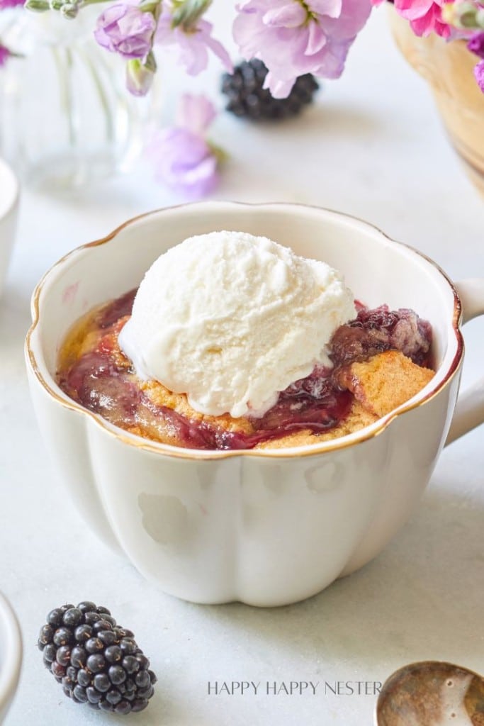 A cup of warm berry cobbler topped with a scoop of vanilla ice cream sits on a white surface. Fresh blackberries and a spoon are nearby, and purple flowers in the background add a decorative touch.